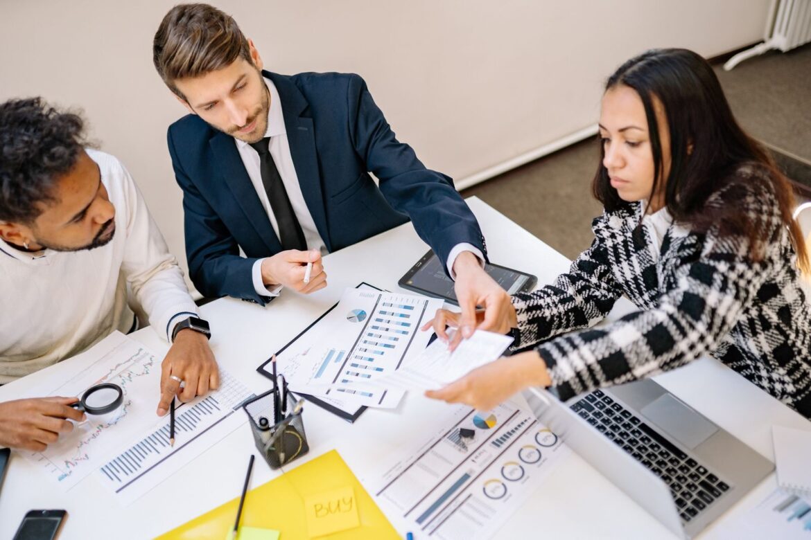 Three colleagues discuss charts and notes around a table.